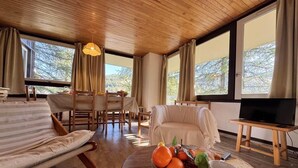 Ein geräumiges Wohnzimmer mit Holzboden, einer Couch, einem Esstisch mit Stühlen, einem Fernseher auf einem Schrank und einer Obstschale auf einem Couchtisch.