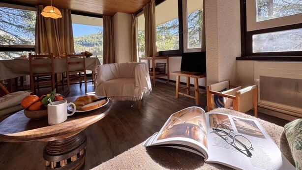 Ein gemütliches Zimmer mit Holzbalkendecke, großen Fenstern, einer Couch, einem Esstisch mit Stühlen und einer kleinen Leseecke mit Zeitschrift und Brillen.