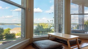 A room with a wooden floor, a small table, and two cushions. There are large windows with a view of a body of water and greenery.