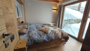 Un lit en bois, recouvert d’une couvre-lit à motifs et agrémenté d’oreillers, une table de chevet et une fenêtre donnant sur un paysage enneigé.
