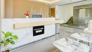 A hotel reception area with a marble counter, a sign indicating it's a company-serviced location, a reception desk, a plant, and a staircase leading to an elevator. - Townhouse Oak Aecs Layout Brookfield (Bengaluru)