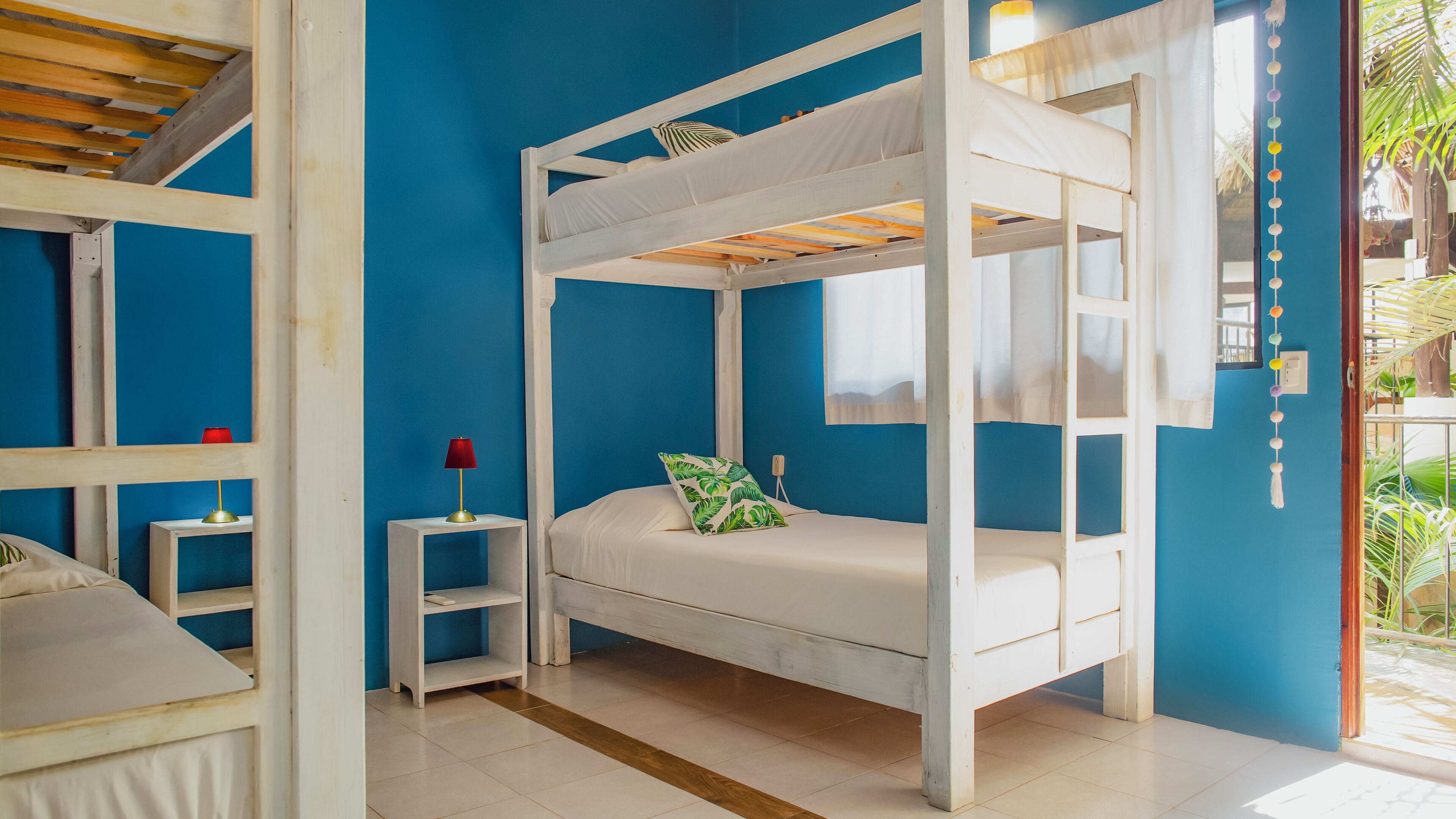 A bunk bed room with a blue wall, a small bedside table with a lamp, and a window with curtains.