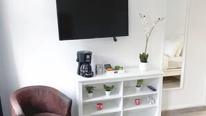 A modern room with a flat-screen TV, a coffee maker, a white shelving unit with plants, a brown leather chair, and a dining table with plates and cutlery.