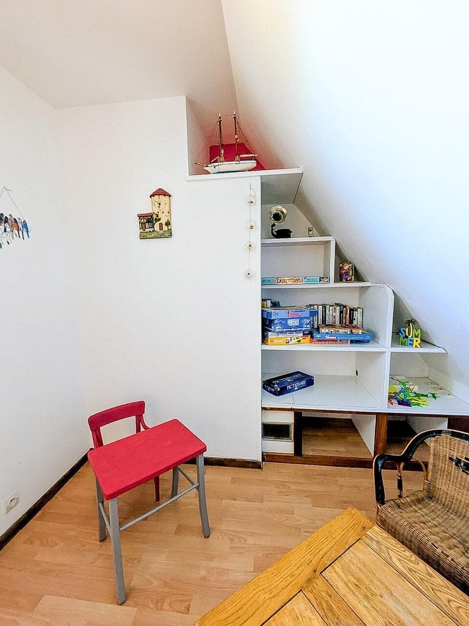 Um quarto com uma cadeira vermelha, uma mesa de madeira, uma estante com livros e jogos, e um pequeno modelo de navio em uma prateleira.