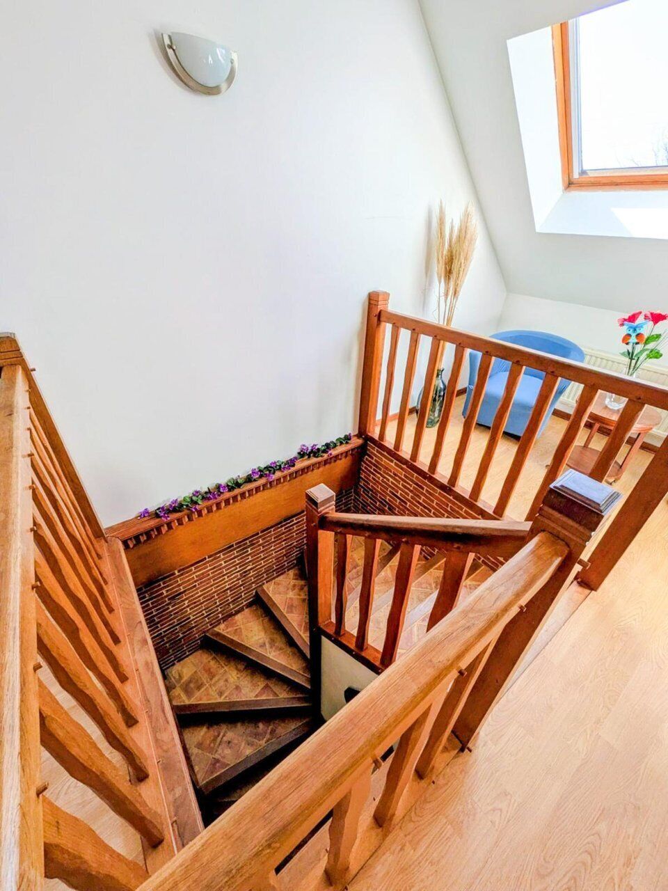 Eine Holztreppe mit Handlauf, ein kleiner Tisch mit Blumen in einer Vase und ein Fenster mit Blick auf den Himmel.