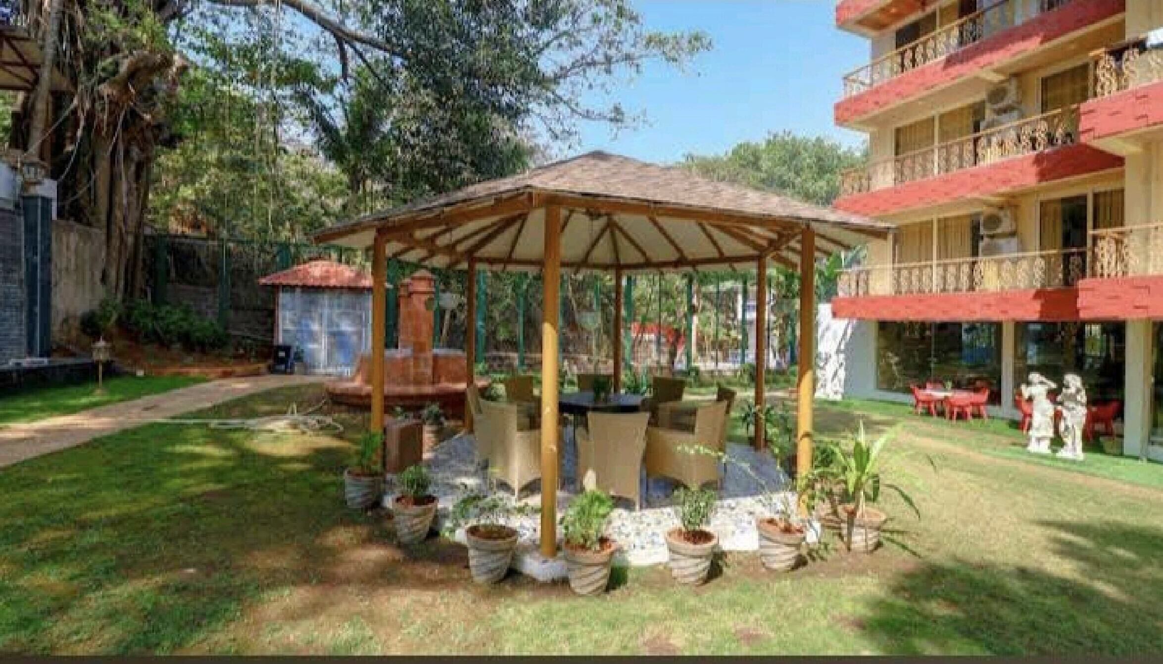 Un área de estar al aire libre cubierta, con mesas y sillas, rodeada de plantas en macetas.