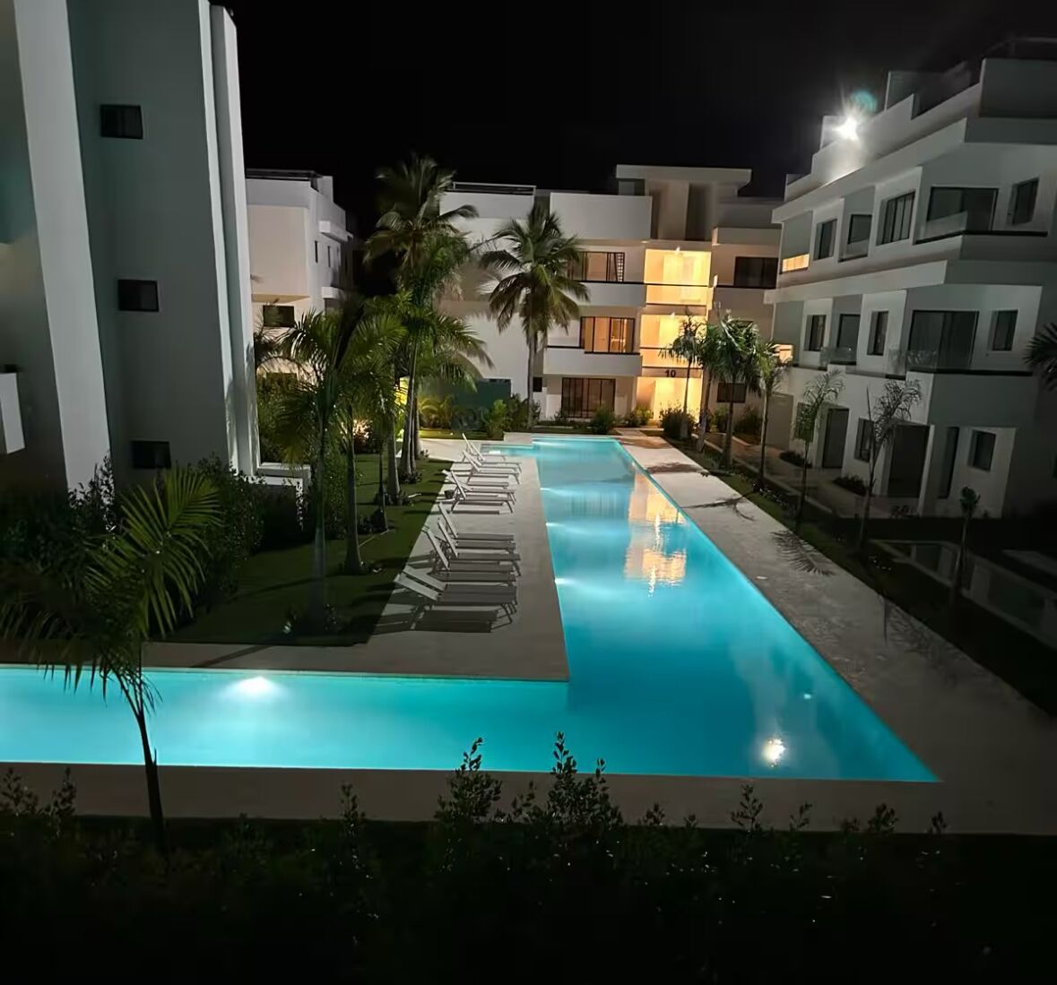 A swimming pool with lounge chairs, palm trees, and modern buildings at night.