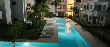 Piscina com espreguiçadeiras, palmeiras e edifícios modernos à noite.