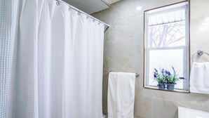 Een badkamer met douchegordijn, handdoekhouder, raam met uitzicht en een potplant.