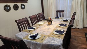 Een eetkamer met een lange tafel gedekt voor zes personen, houten stoelen, een raam met planten en een gordijn.