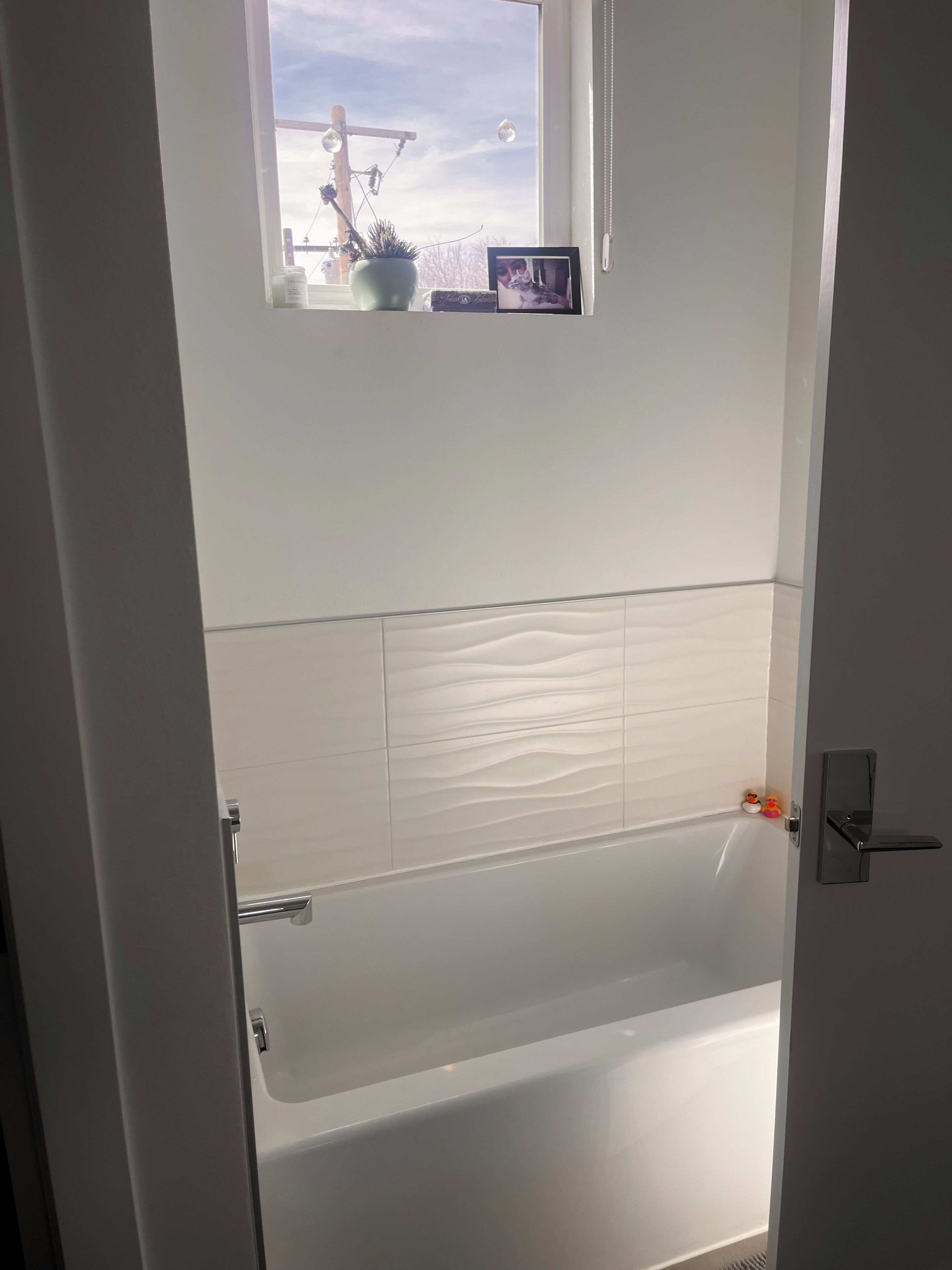 A bathroom with a bathtub, a window with a view of the outside, and a small shelf with a picture frame and a plant.