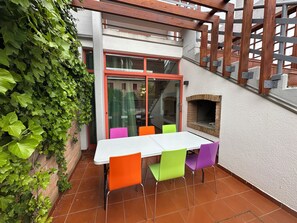 Eine Terrasse mit einem weißen Tisch, bunten Stühlen, einem Backofen aus Ziegeln und einer Holzpergola.