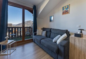 Ein modernes Wohnzimmer mit einer grauen Couch, einem hölzernen Beistelltisch und Blick auf Berge durch eine Glastür.