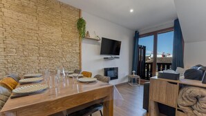 Una sala de estar moderna con una pared de piedra, un televisor de pantalla plana, una mesa de comedor dispuesta con platos y vasos, un sofá y vistas de un paisaje urbano a través de ventanas amplias.