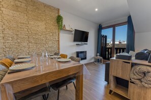 Ein modernes Wohnzimmer mit Steinwand, Flachbildfernseher, Esstisch gedeckt mit Tellern und Gläsern, einer Couch und Blick auf eine Stadtansicht durch große Fenster.