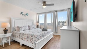 Ein Schlafzimmer mit Bett, Nachttisch, Lampe, Deckenventilator, Fernseher und einem Fenster mit Vorhängen.