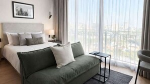 Ein modernes Hotelzimmer mit einem Bett, einem grünen Sofa, einem kleinen Tisch, einem Deckenventilator und großen Fenstern mit Blick auf die Stadt.