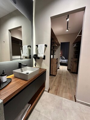 Un bagno moderno con uno specchio grande, un lavandino bianco e un mobile vanitario in legno. C'è un porta asciugamani con due asciugamani, una bottiglia di sapone per le mani e un telefono nero. Il bagno dà accesso a una camera da letto con un letto e un armadio.