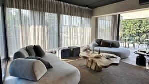Ett modernt vardagsrum med en böjd soffa, ett runt soffbord och ett stort fönster med tunna, genomskinliga gardiner.