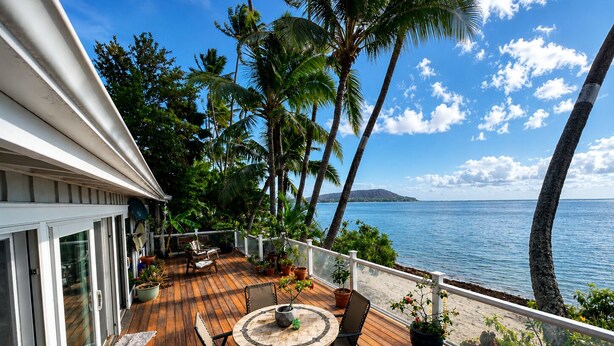 Eine Holzterrasse mit rundem Tisch und Stühlen, umgeben von Kübelpflanzen, mit Blick auf einen Strand mit Palmen und den Ozean.
