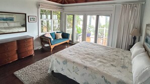 Ein Schlafzimmer mit Bett, Schminktisch, Sessel, Deckenventilator und großen Fenstern.