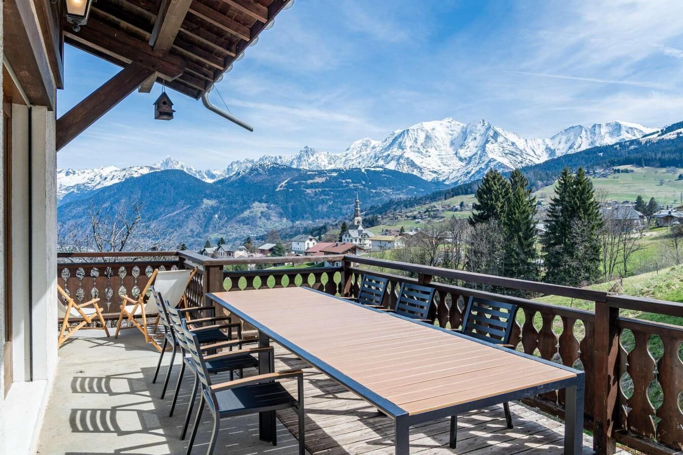 A balcony with a wooden table and chairs, overlooking a mountainous landscape with snow-capped peaks and a village below.