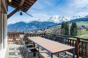Ein Balkon mit Holztisch und -stühlen, mit Blick auf eine Berglandschaft mit schneebedeckten Gipfeln und einem Dorf darunter.