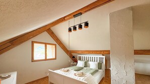 A bedroom with a sloped ceiling, a bed with white headboard, a wooden floor, and a window with a view.