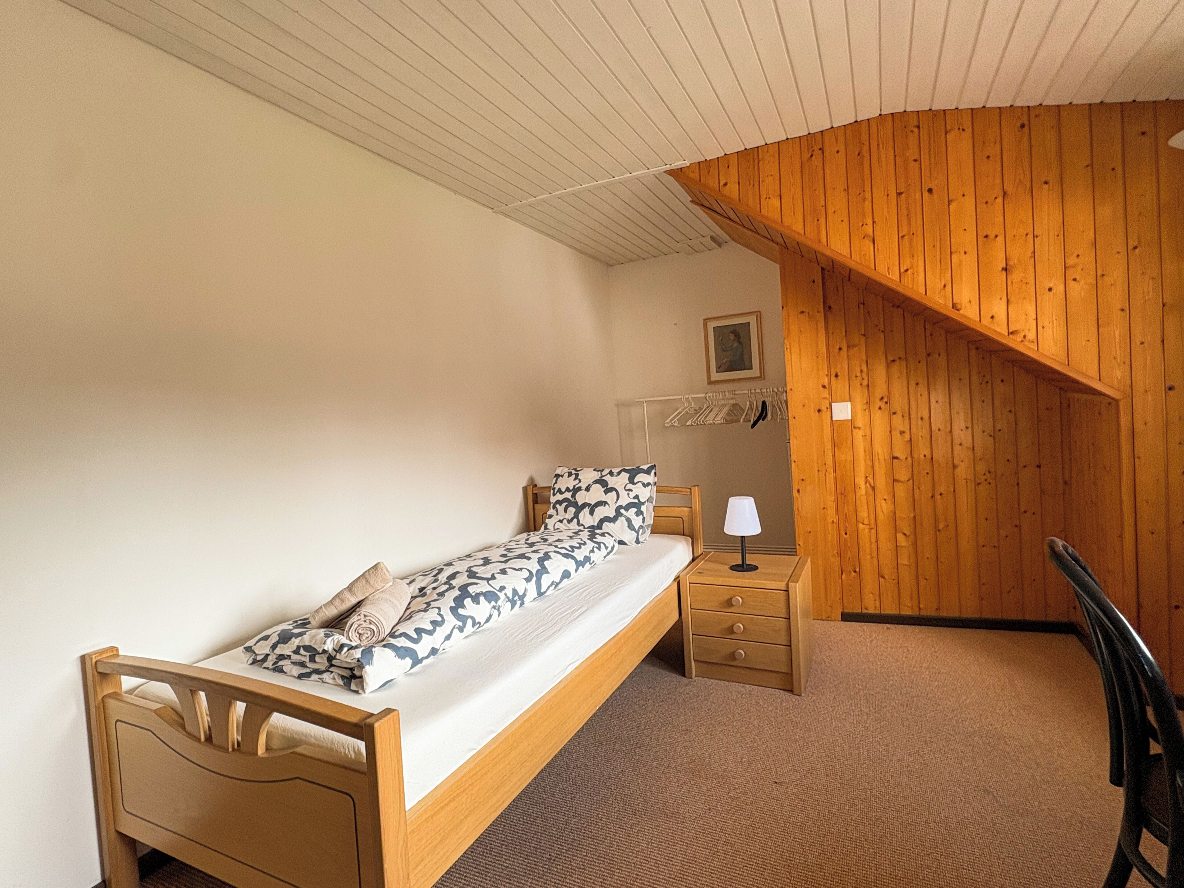 Ein Schlafzimmer mit einem Bett, einem Nachttisch, einer Lampe, einem Sessel und einer Holzbalkendecke.