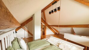 A bedroom with a sloped ceiling, wooden beams, a bed with green bedding, a white dresser, and a hanging light fixture.
