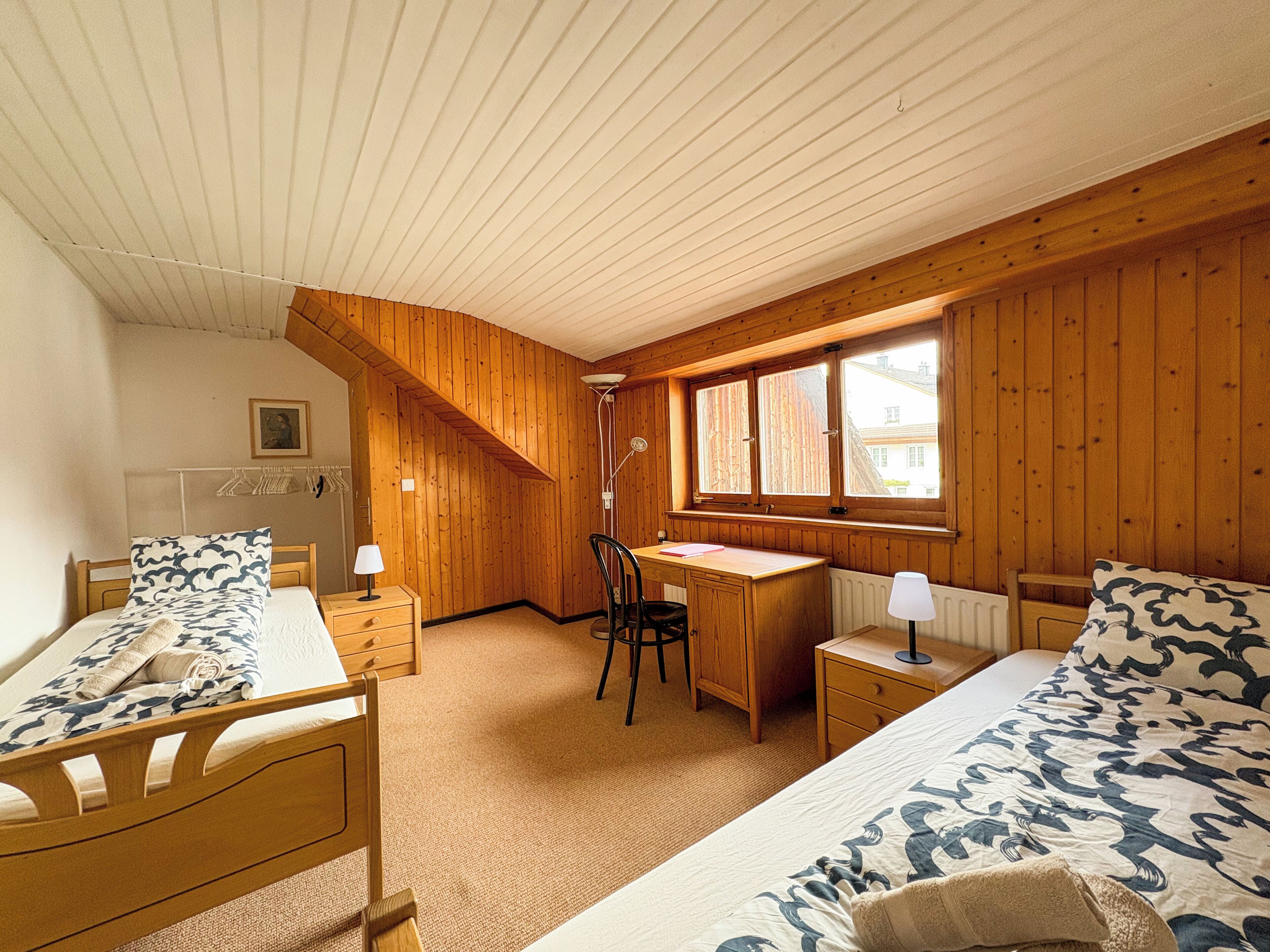 A bedroom with a sloped ceiling, wooden walls, a desk, two beds, and a chair.