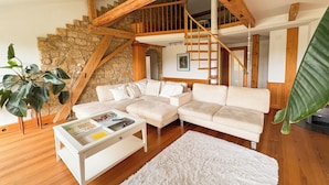 A modern living room with a white sofa, a glass coffee table, a white rug, wooden flooring, a stone wall, and a wooden staircase.