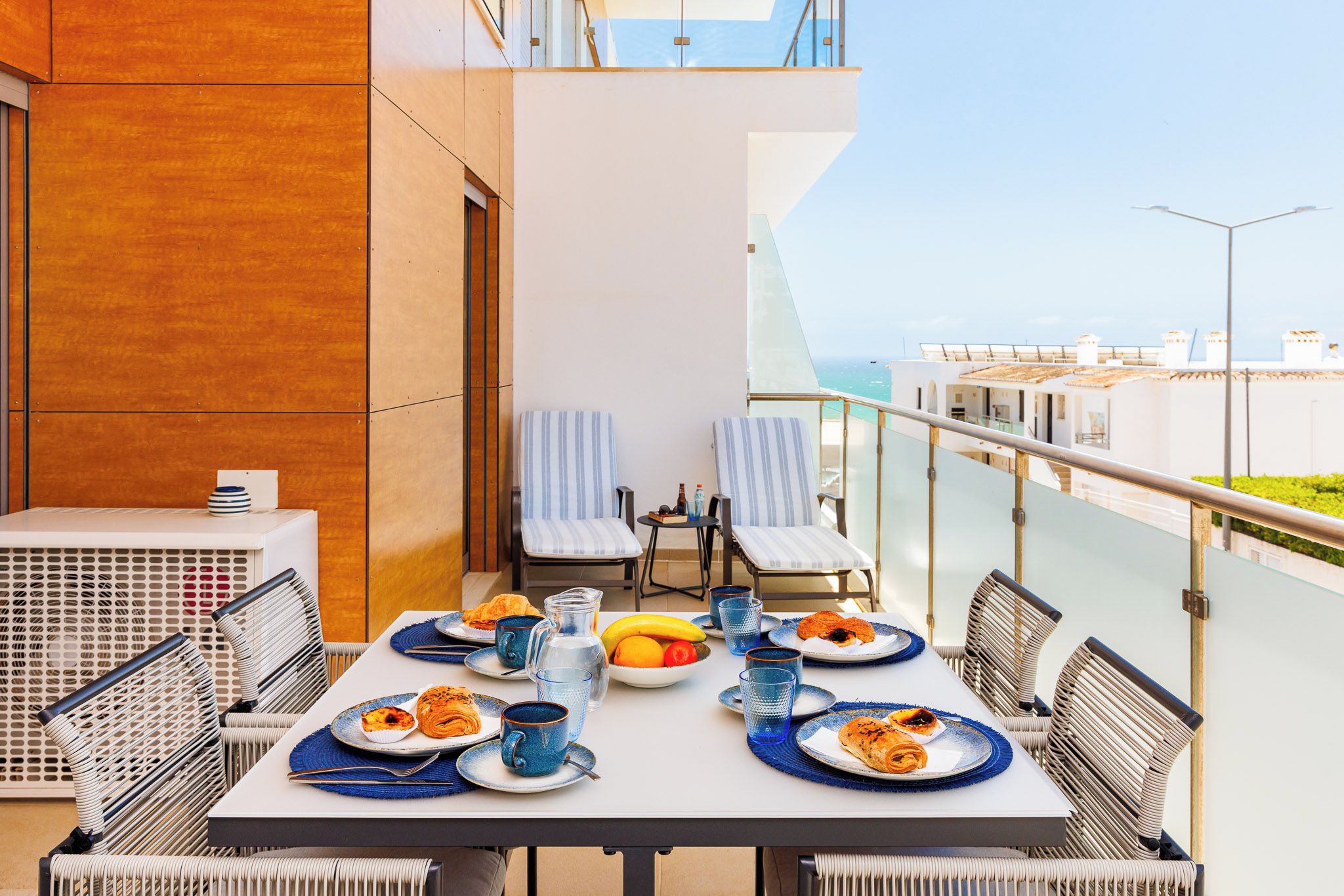A dining table set with plates, glasses, and a fruit bowl on a balcony with a view of the sea and buildings.
