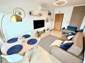 A modern living room with a round table, sofa, and TV. - Beachside 1BR Stay for 4 – Easy Beach Access by Lift & Free Parking (Olhos de Água)