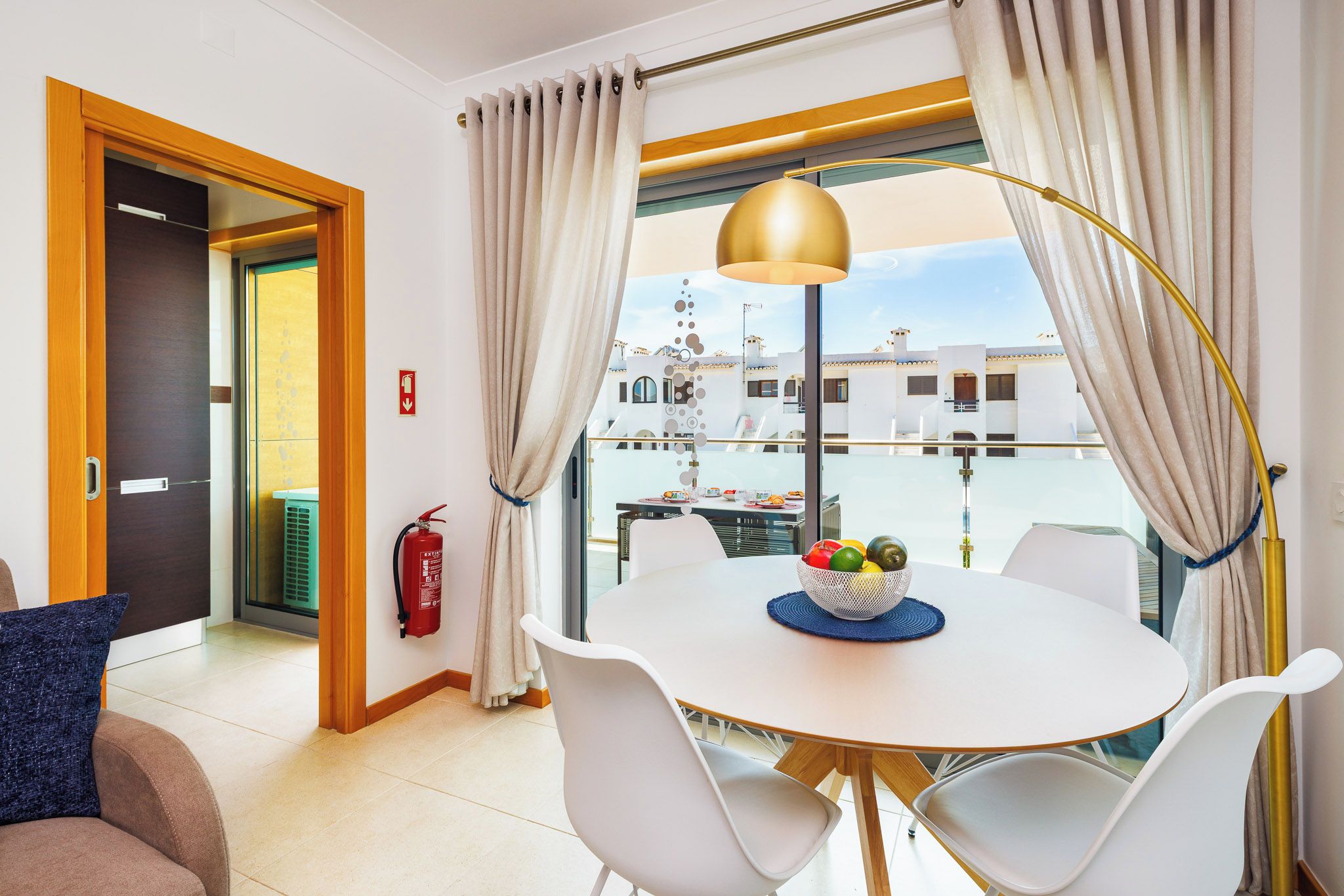 A modern dining area with a round table, white chairs, a fruit bowl, and a view of buildings through large windows.