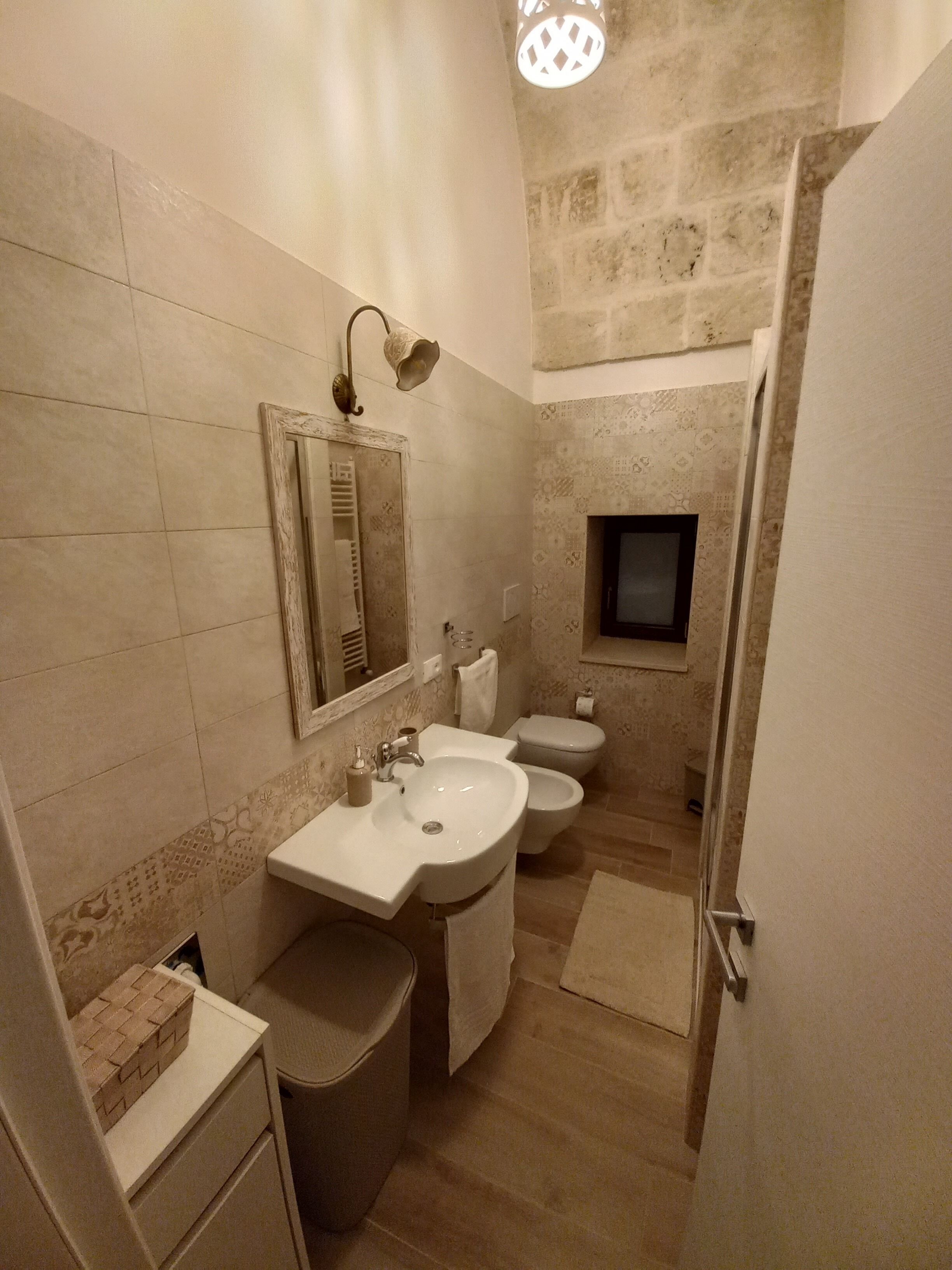A bathroom with a sink, toilet, and bidet, tiled walls, and a small window.
