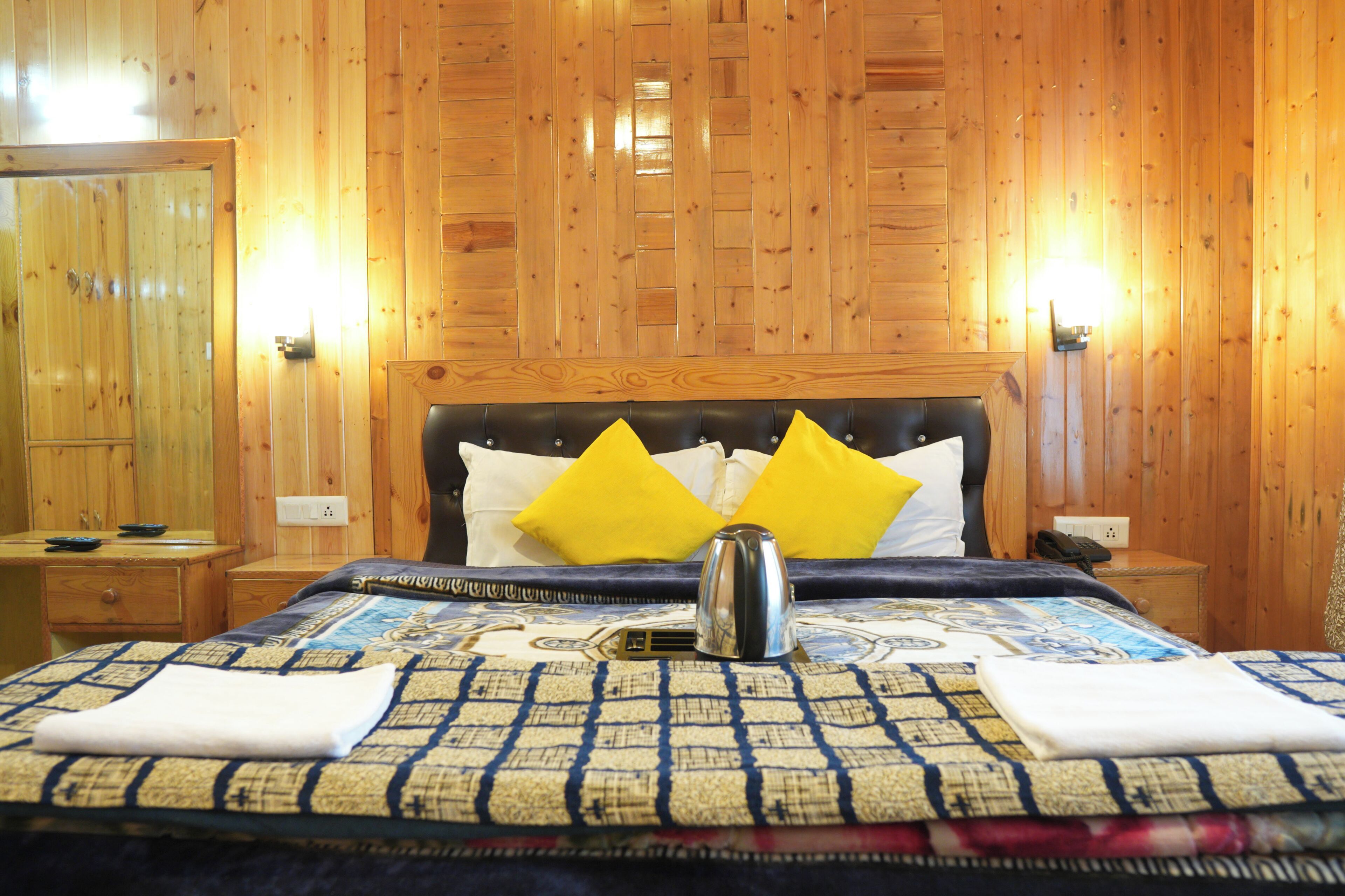 A wooden-paneled room with a bed, two yellow pillows, a kettle, and a quilted bedspread.