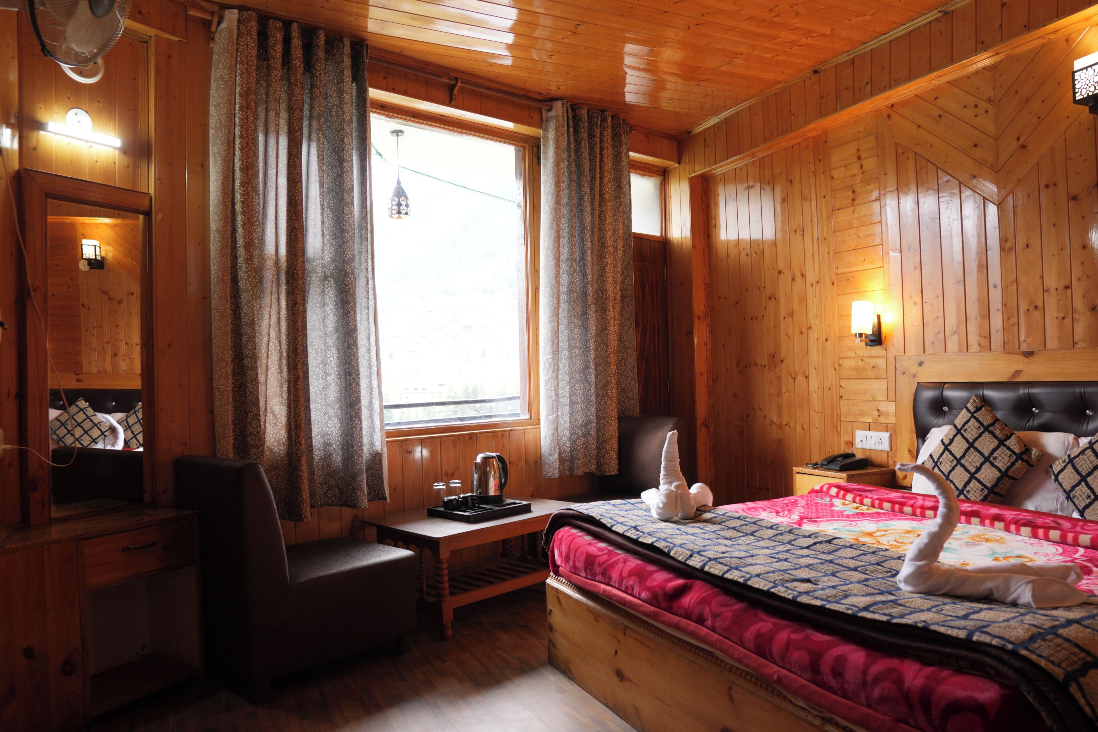 A wooden cabin room with a bed, desk, chair, and a window with curtains.