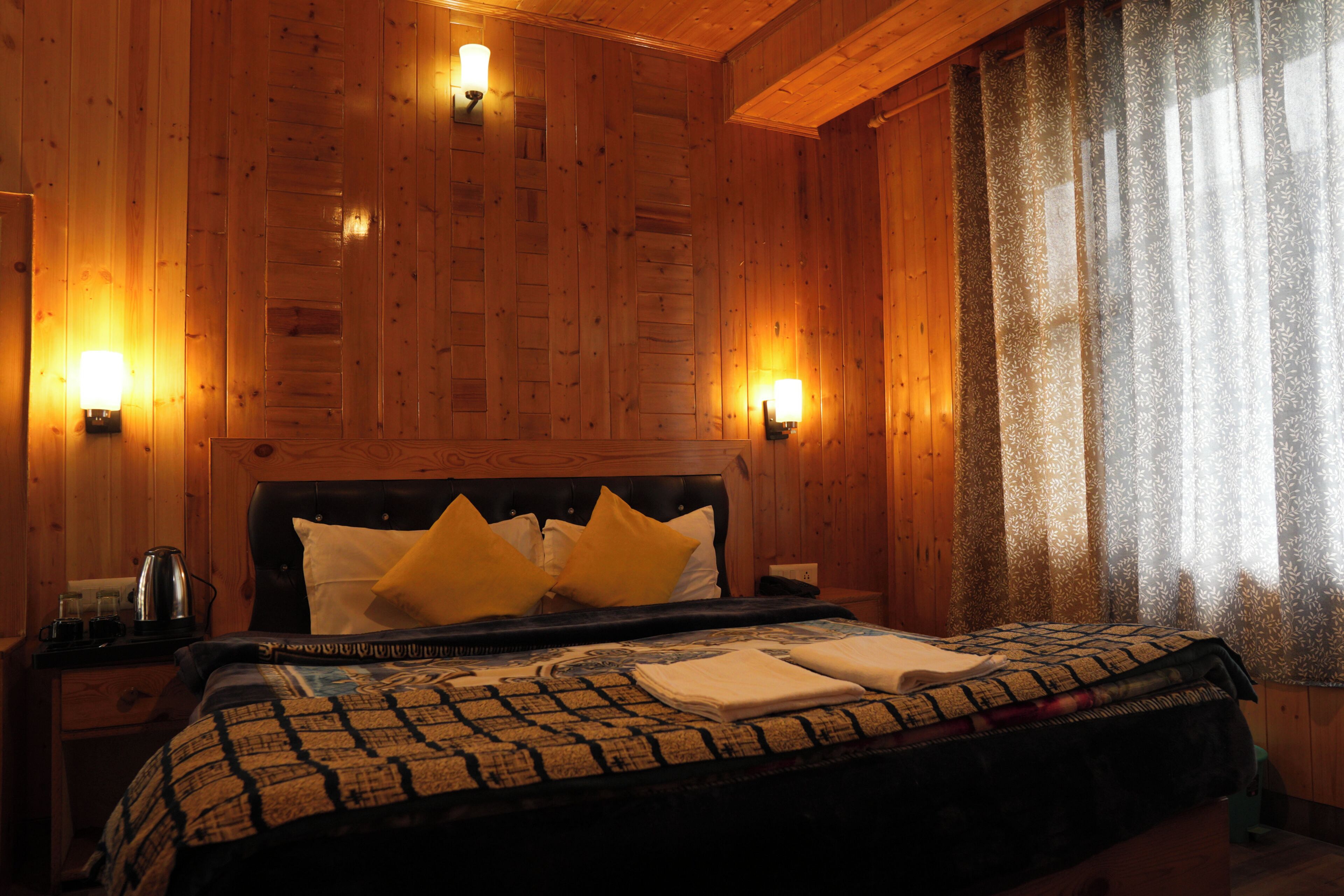 A wooden-paneled room with a bed, two bedside lamps, and a window with patterned curtains.