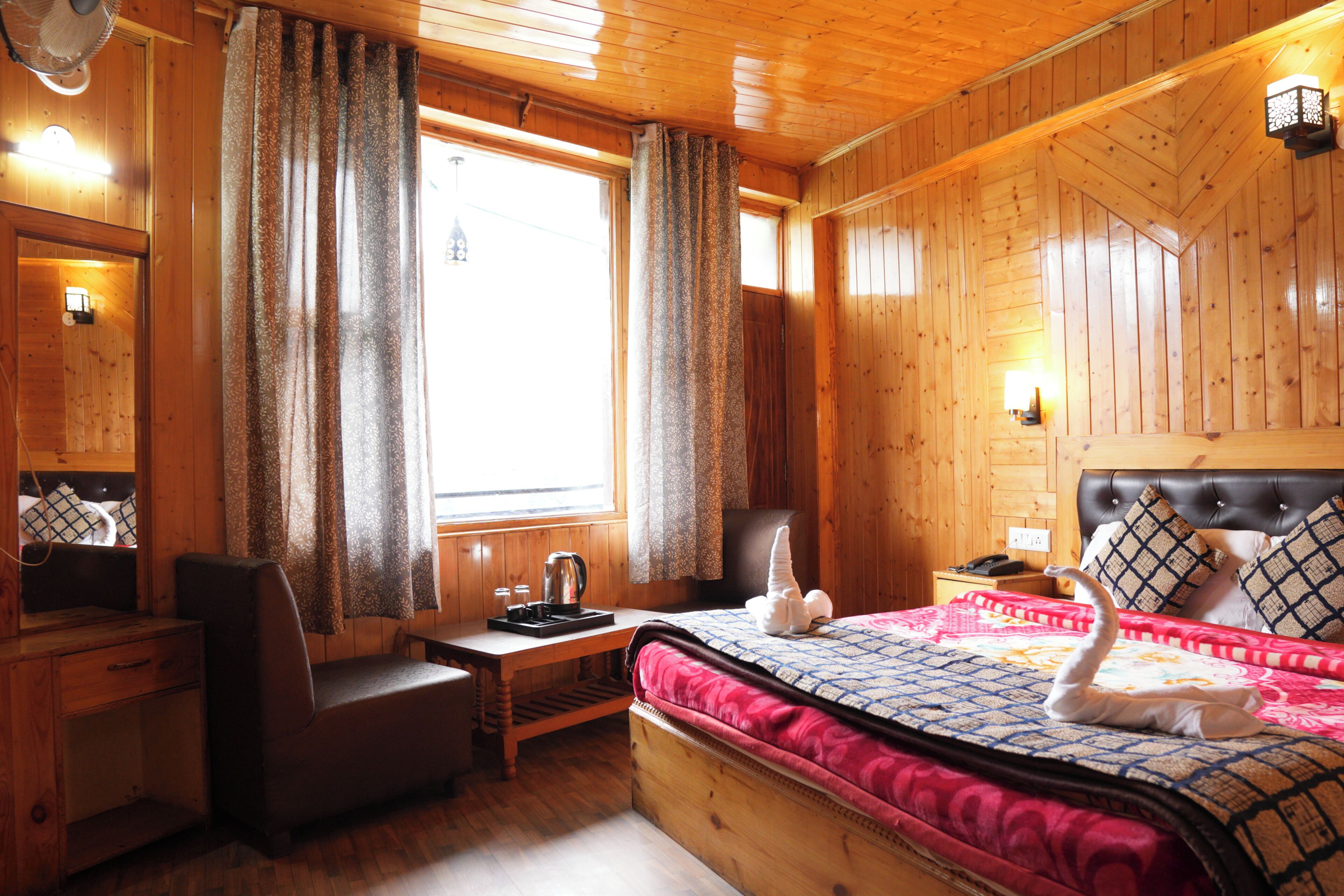 A wooden cabin room with a bed, a desk, a chair, a window with curtains, and a wall-mounted lamp.