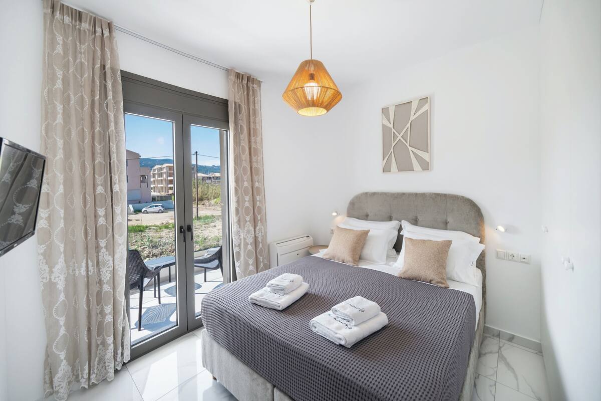Una camera da letto con un letto, tende, un lampadario a sospensione, una TV a parete e vista attraverso una porta in vetro.
