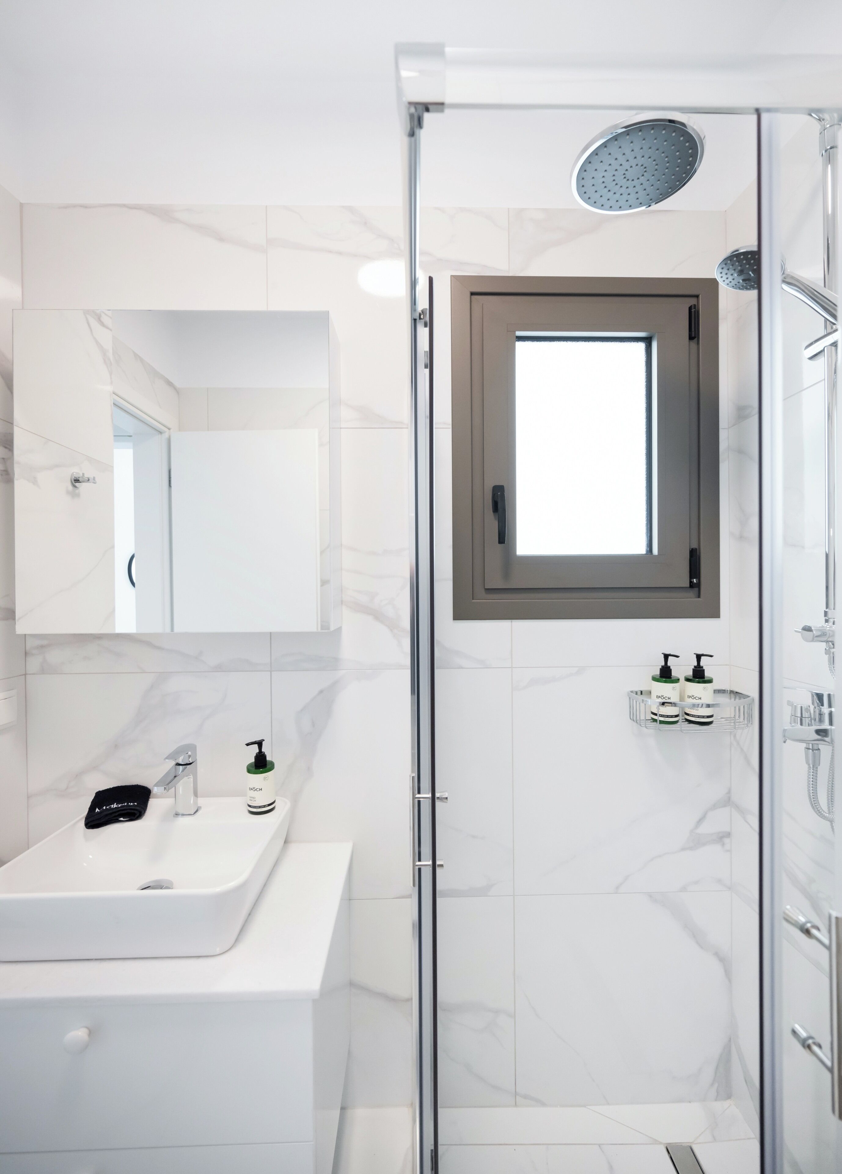 A modern bathroom with a glass shower enclosure, a white sink with a mirror, and a small window.