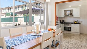 A dining area with a table set for four, a kitchen with white cabinets and appliances, and large windows offering a view of the outside. - Pou de s'Alou by Rentallorca (Sineu)