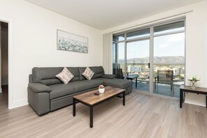A modern living room with a grey sofa, a wooden coffee table, and a large window offering a city view.