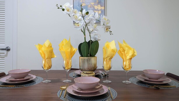 A dining table set with pink plates, bowls, and glasses, accompanied by a vase of white orchids and yellow napkins. - 1BR Universal Studios Pool Gym Parking (North Hollywood)