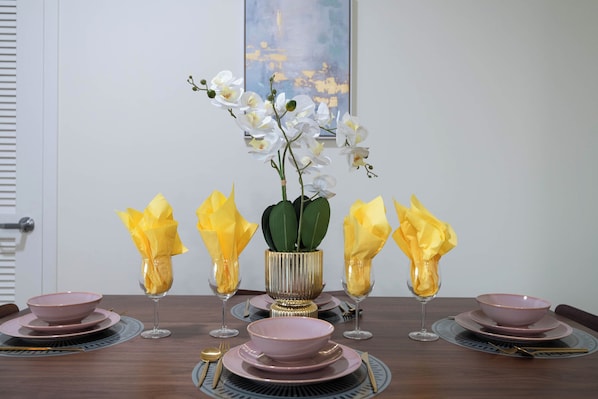 Une table à manger dressée avec de l’argenterie rose, des assiettes, des bols et des verres, agrémentée d’un vase d’orchidées blanches et de serviettes jaunes.