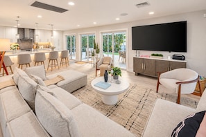 A modern living room with a large sofa, a coffee table, and a television. There is a kitchen area with bar stools and a dining table visible through the glass doors. - Direct Lake Ida Access | Sleeps 8 | Golf Cart | Heated Pool | by Delray Nightly (Boynton Beach)