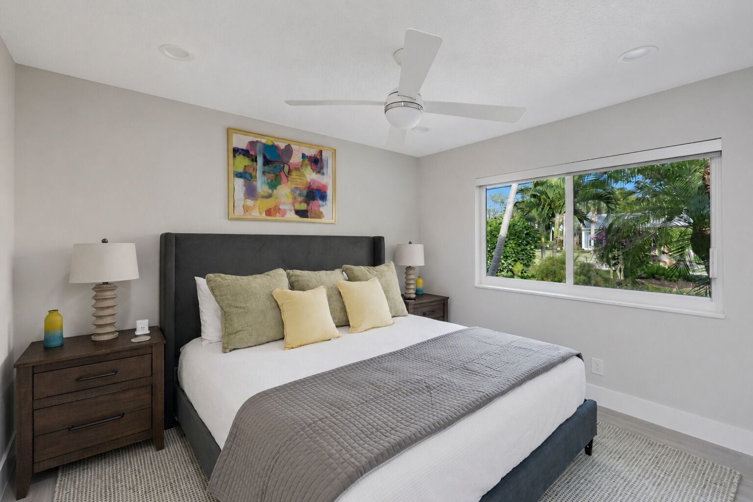A bedroom with a bed, nightstands, a ceiling fan, a window with a view of greenery, and a colorful abstract painting on the wall.