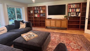 Ein Wohnzimmer mit einer Couch, einem Hocker, einem Bücherregal, einem Flachbildfernseher und einem großen Fenster.