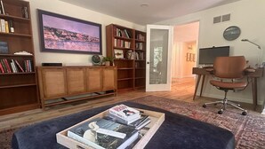 Ein Wohnzimmer mit einem dunkelblauen Ottomanen, Holzbücherregalen, einem Fernseher und einem Schreibtisch mit einem Stuhl.